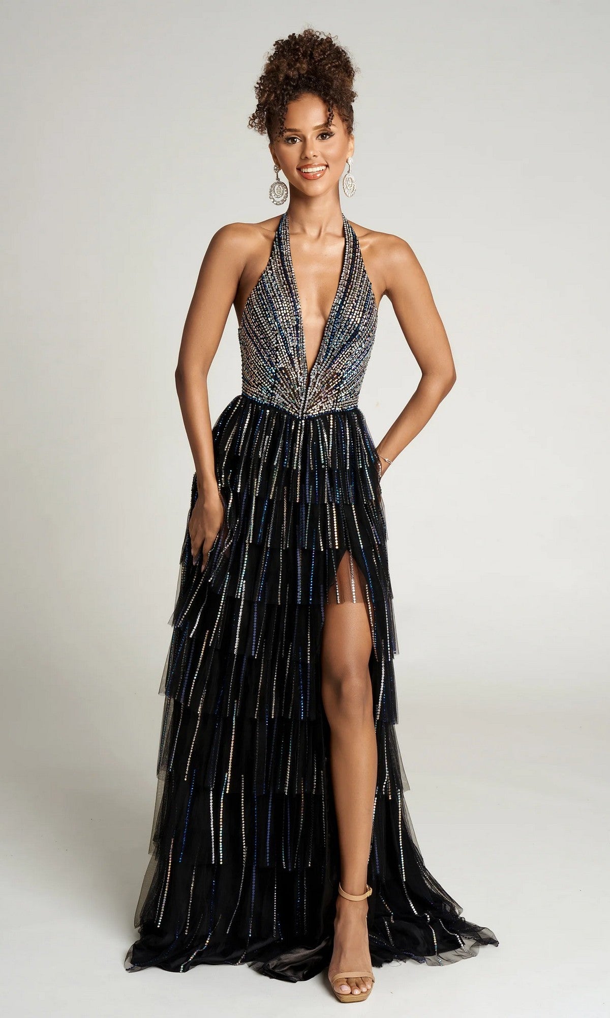 Woman standing and smiling in the Long Formal Dress: Portia and Scarlett PS26117—a sleeveless, deep V-neck black gown with beadwork, tiered shimmering layers, high slit—accessorized with hoop earrings and heels.